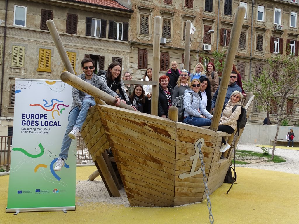 People are posing in a toy wooden boat, with a roll up of Europe Goes Local to the left